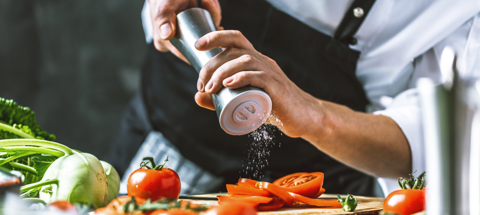 Koch hält mit beiden Händen eine Salzmühle und streut damit Salz auf Tomaten